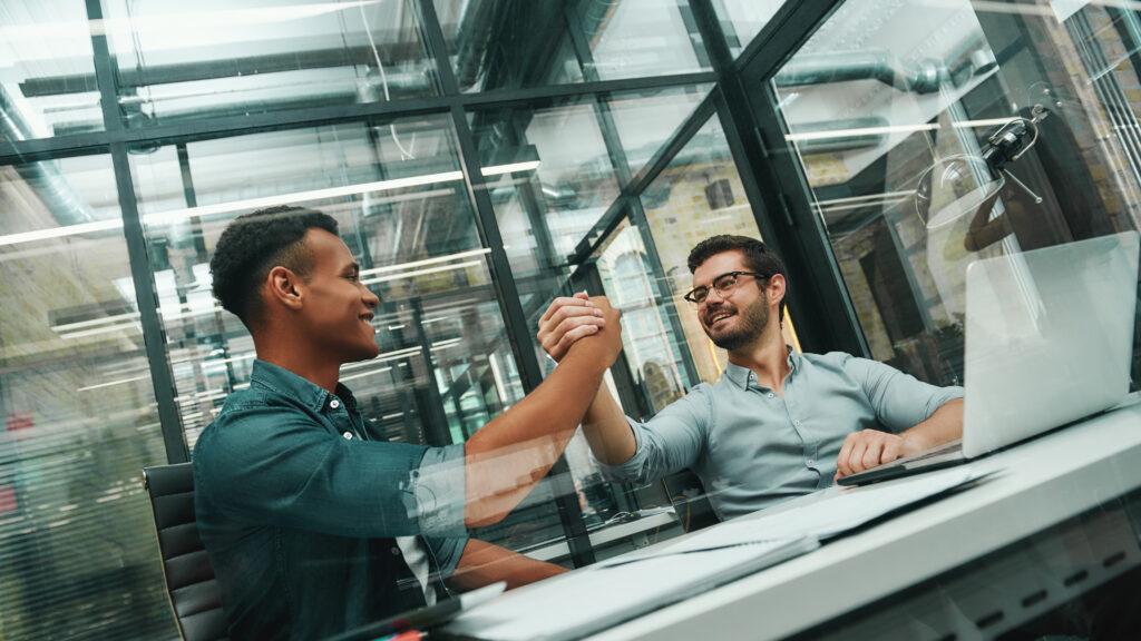 Glad to work with you Two young and handsome men shaking hands and smiling while working in modern office